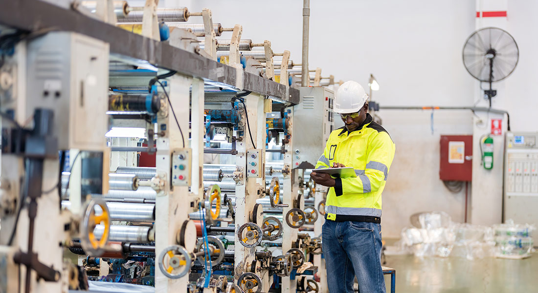 A machinery worker is inspecting the workspace with a clipboard. He is wearing a hard hat, a yellow shirt, and safety glasses.