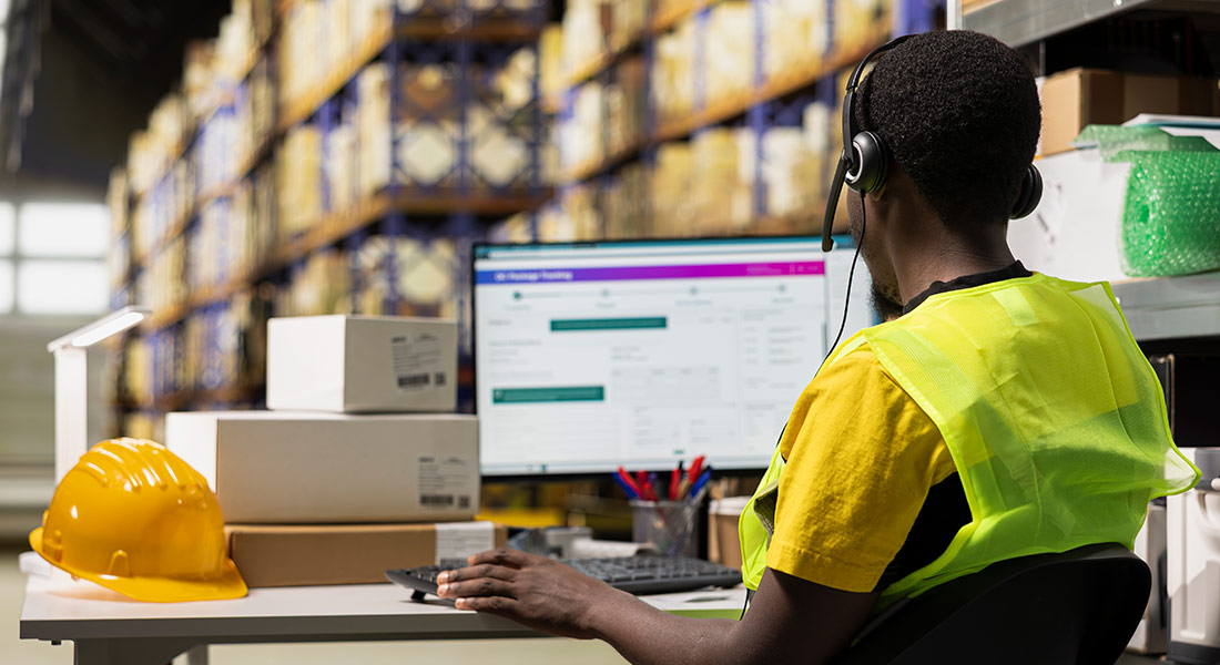 a warehouse worker wearing a headset is looking at inventory on a computer; there are shipping boxes on the workstation table