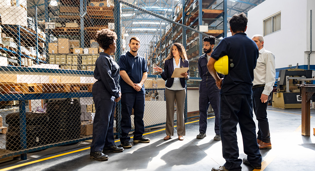 A warehouse manager is meeting with the company's team members in a warehouse filled with inventory.