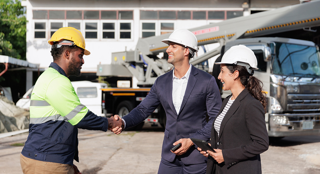 A construction engineer shaking hands with an equipment vendor sales manager. Large commercial crane equipment is in the background.